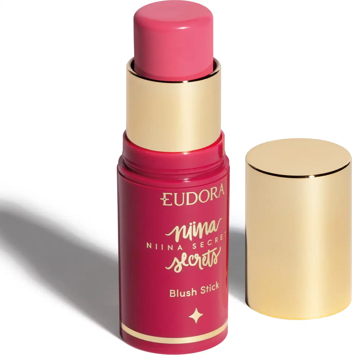O produto apresentado é um blush stick da linha Niina Secrets da marca Eudora. Possui uma embalagem rosa com detalhes dourados, incluindo o nome da marca e da linha. O produto tem formato cilíndrico e tampa dourada.
