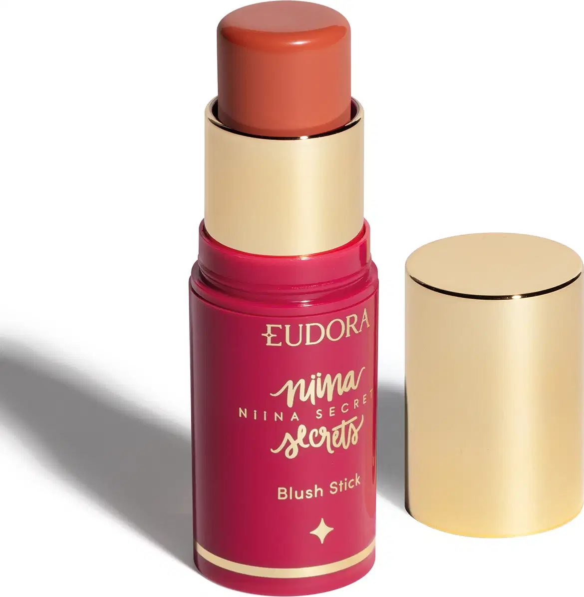 O produto apresentado é um blush stick da linha Niina Secrets da marca Eudora. Possui uma embalagem rosa com detalhes dourados, incluindo o nome da marca e da linha. O produto tem formato cilíndrico e tampa dourada.