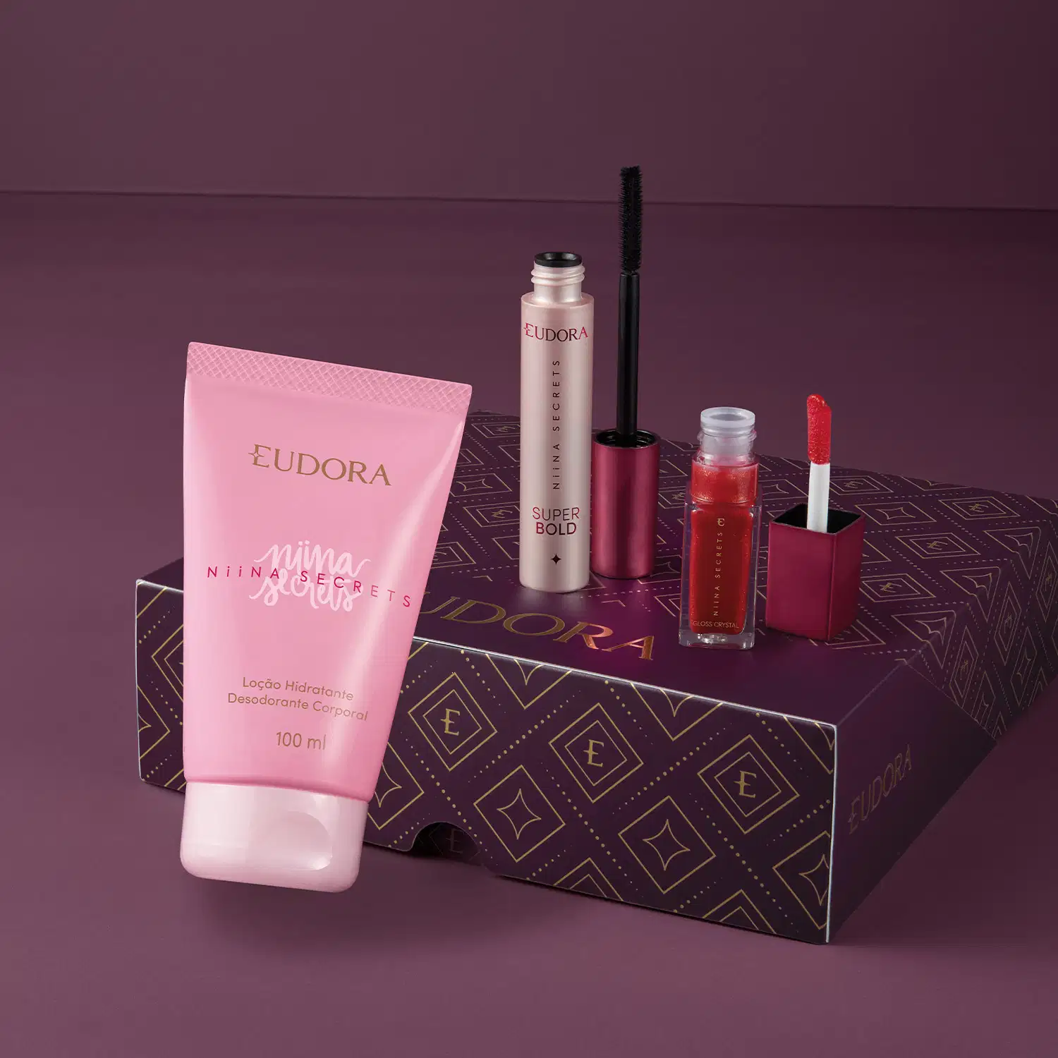 A caixa roxa e dourada da marca Eudora contém diversos produtos de beleza, incluindo uma loção hidratante corporal da linha Nina Secrets, um gloss que está aberto e um frasco de máscara de cílios preta também com o aplicador ao lado.