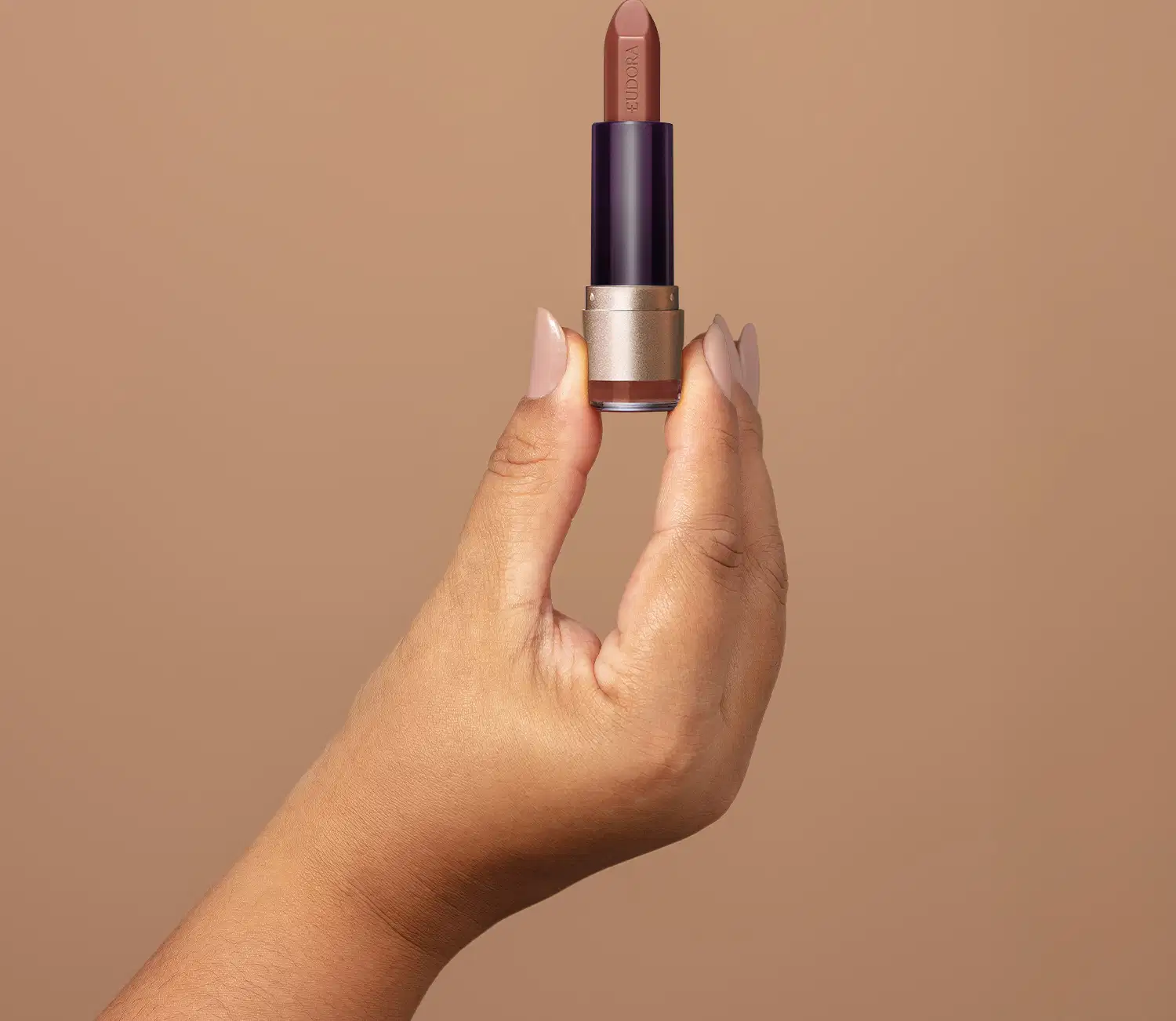 Imagem de um batom da marca Eudora em um fundo bege, com uma mão segurando o produto. O batom possui uma embalagem escura e uma ponta nude, sugerindo um tom de cor natural e sutil para uso diário.