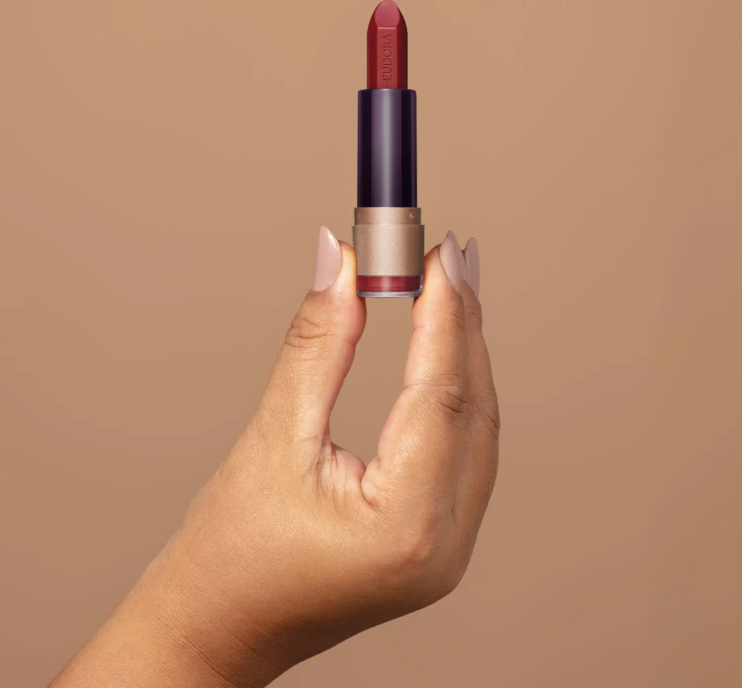 Embalagem de batom vermelho da marca Eudora Make sobre superfície bege, com mão segurando o produto em destaque.