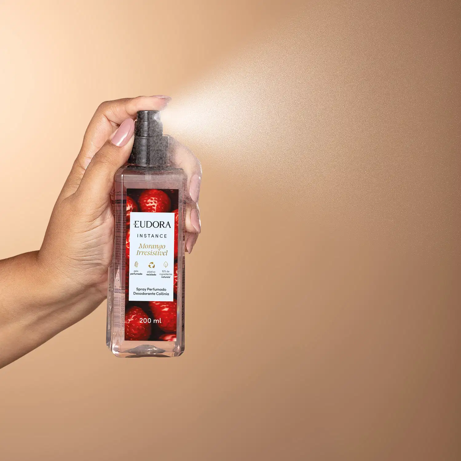 O produto mostrado é um spray perfumado da marca Eudora, que está sendo segurado por uma mão feminina. O borrifador está acionado, saindo o produto em forma de spray. O frasco é transparente com uma tampa preta e um rótulo vermelho e branco.