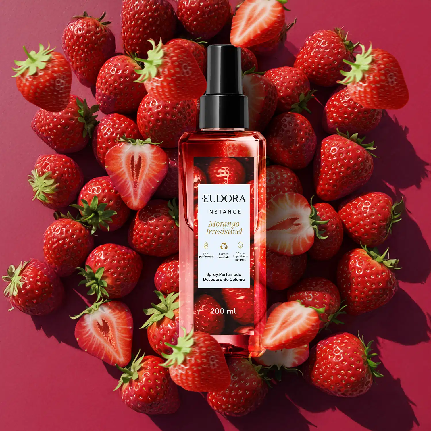 Imagem ambientada do Spray Perfumado da marca Eudora, da linha Instance Morango. O frasco é transparente com um borrifador preto e um rótulo vermelho e branco. Abaixo do produto temos vários morangos dispostos em um fundo vermelho bordô.
