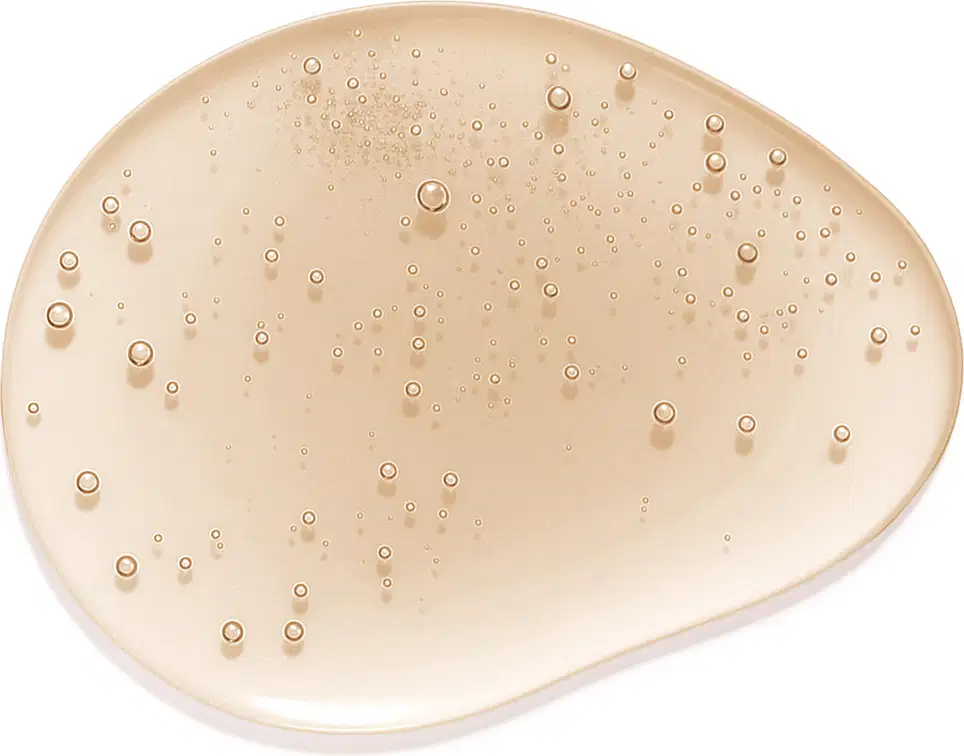 A superfície do produto apresenta uma textura suave e cremosa, com uma tonalidade bege clara e uma aparência levemente brilhante, com pequenas bolhas dispersas por toda a superfície, sugerindo uma consistência leve.