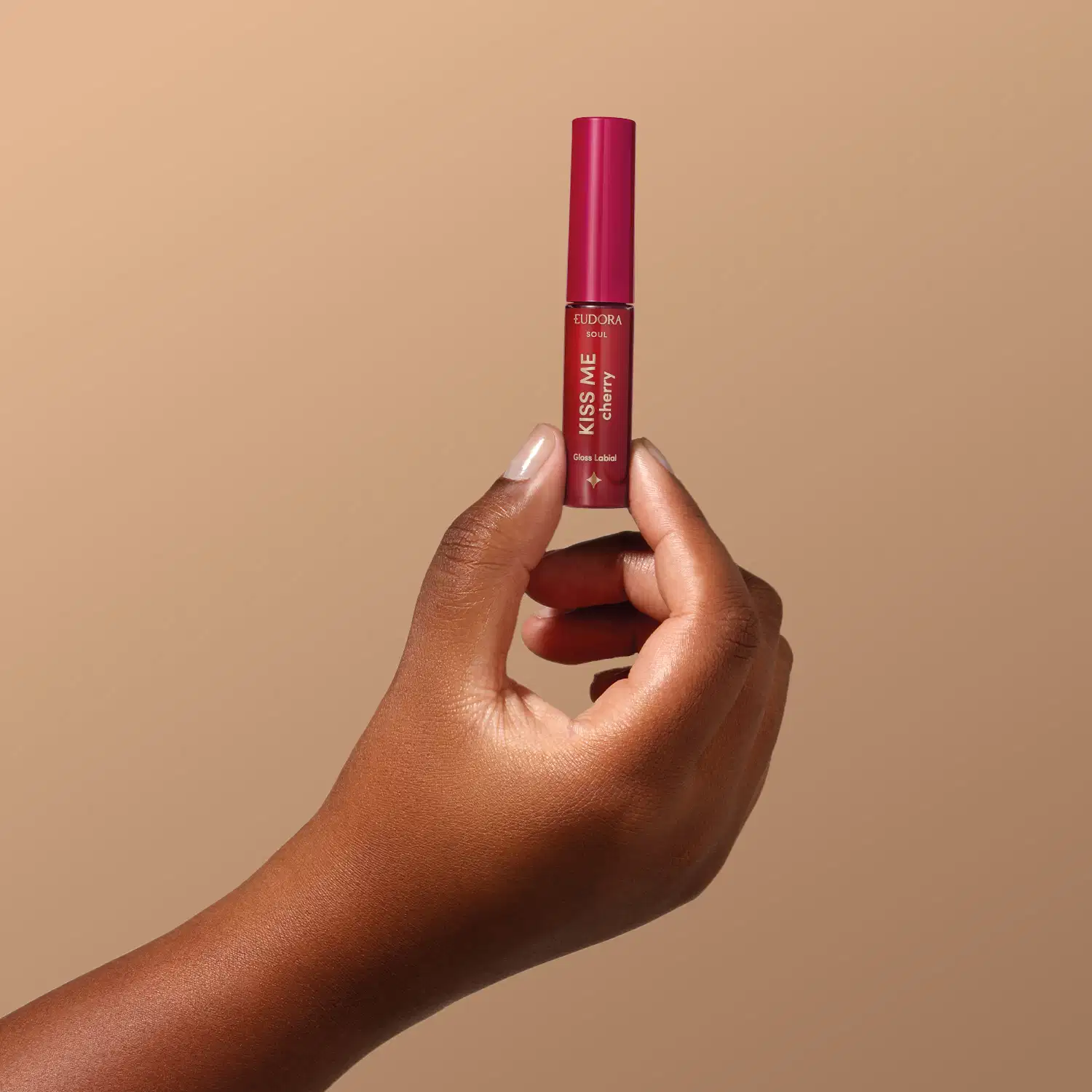 O produto mostrado está sendo segurado por uma mão, é o Gloss Labial da marca Eudora, da linha Kiss Me. Possui uma embalagem cilíndrica de cor vermelha, com o nome da linha "Kiss Me".