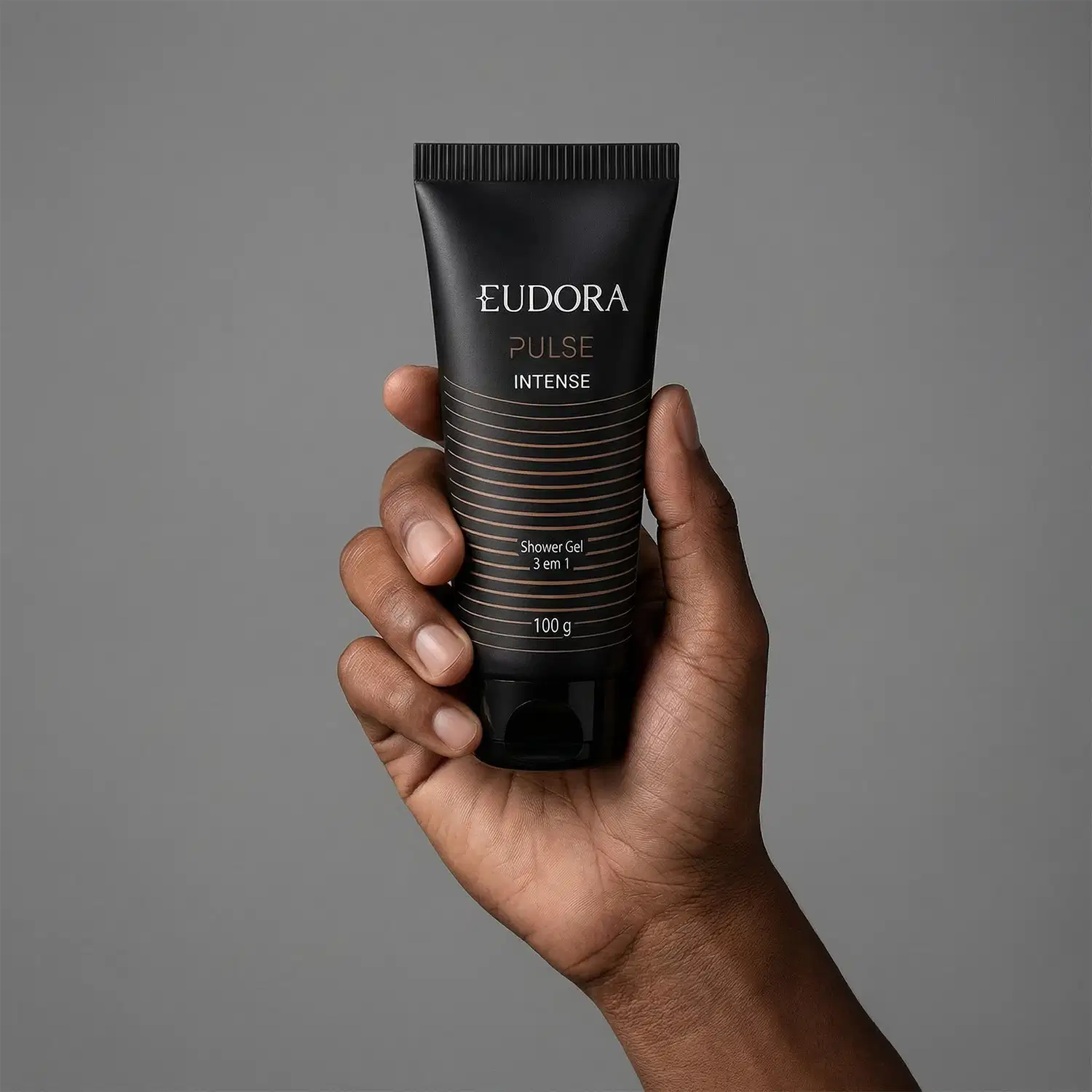 Embalagem preta e dourada do produto shower gel da marca Eudora, em um ambiente minimalista e monocromático, sendo segurado por uma mão masculina.