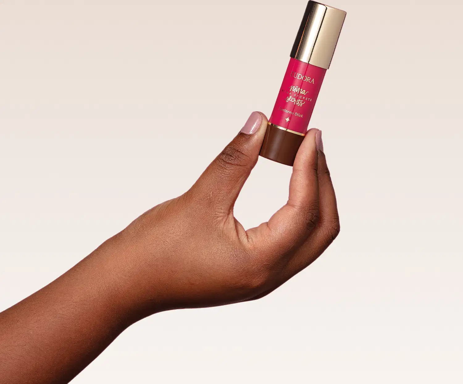 O produto mostrado na imagem é o Contorno Stick de Niina Secrets, que possui uma embalagem cilíndrica dourada com um rótulo rosa brilhante. O produto está sendo segurado por uma mão com pele escura.