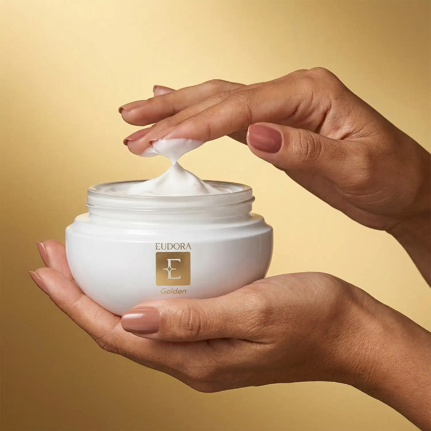 Mãos segurando um pote de creme da marca Eudora Golden em um fundo bege/dourado, sugerindo um ambiente de cuidados de beleza e cosmética.