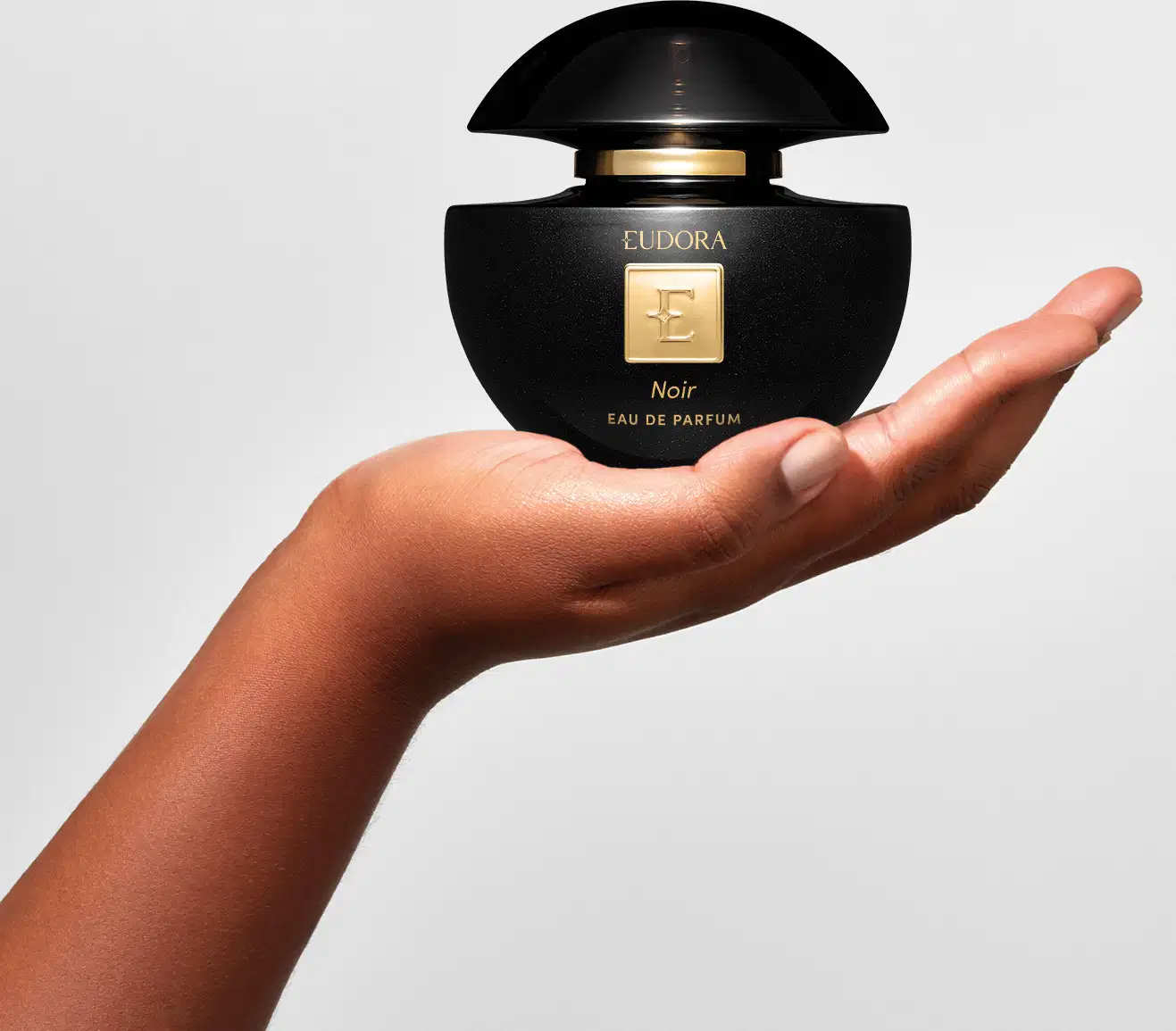 Mão feminina segurando um frasco de Eudora Noir Eau de Parfum preto e dourado da marca Eudora, em um ambiente claro e minimalista, sugerindo um contexto de beleza e sofisticação.