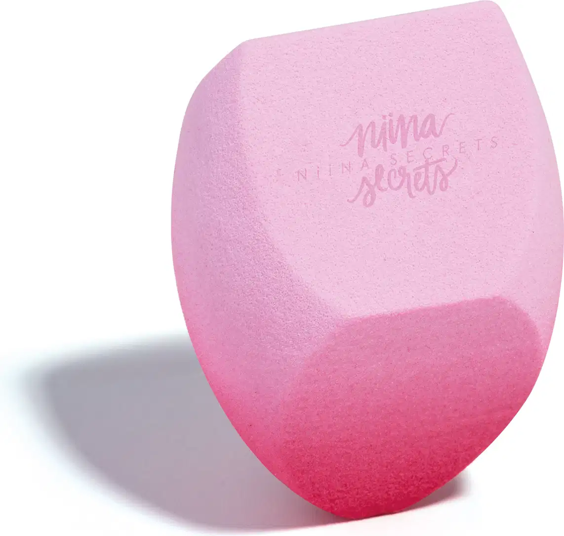 O produto apresentado é uma esponja para maquiagem da marca Niina Secrets, com uma coloração degradê de rosa claro a rosa escuro. Possui um formato arredondado e achatado em diferentes partes. Em outro ângulo.