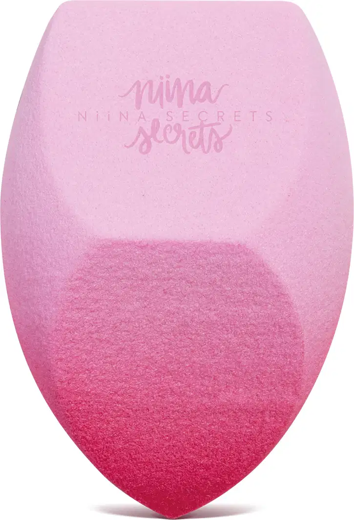 O produto apresentado é uma esponja para maquiagem da marca Niina Secrets, com uma coloração degradê de rosa claro a rosa escuro. Possui um formato arredondado e achatado em diferentes partes.