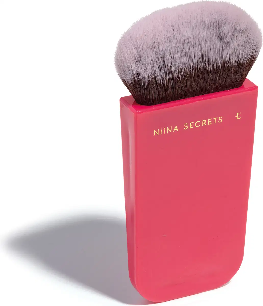 O produto apresentado é um pincel de maquiagem da marca Niina Secrets. O pincel possui cerdas macias e uma base em um tom rosa vibrante, com o nome da marca impresso em letras douradas. Em outro ângulo.