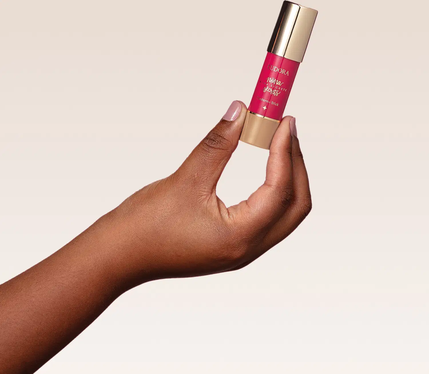 O produto mostrado na imagem é o Contorno Stick de Niina Secrets, que possui uma embalagem cilíndrica dourada com um rótulo rosa brilhante. O produto está sendo segurado por uma mão com pele escura.