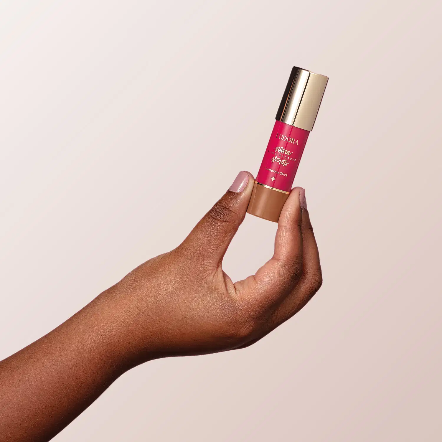 O produto mostrado na imagem é o Contorno Stick de Niina Secrets, que possui uma embalagem cilíndrica dourada com um rótulo rosa brilhante. O produto está sendo segurado por uma mão com pele escura.
