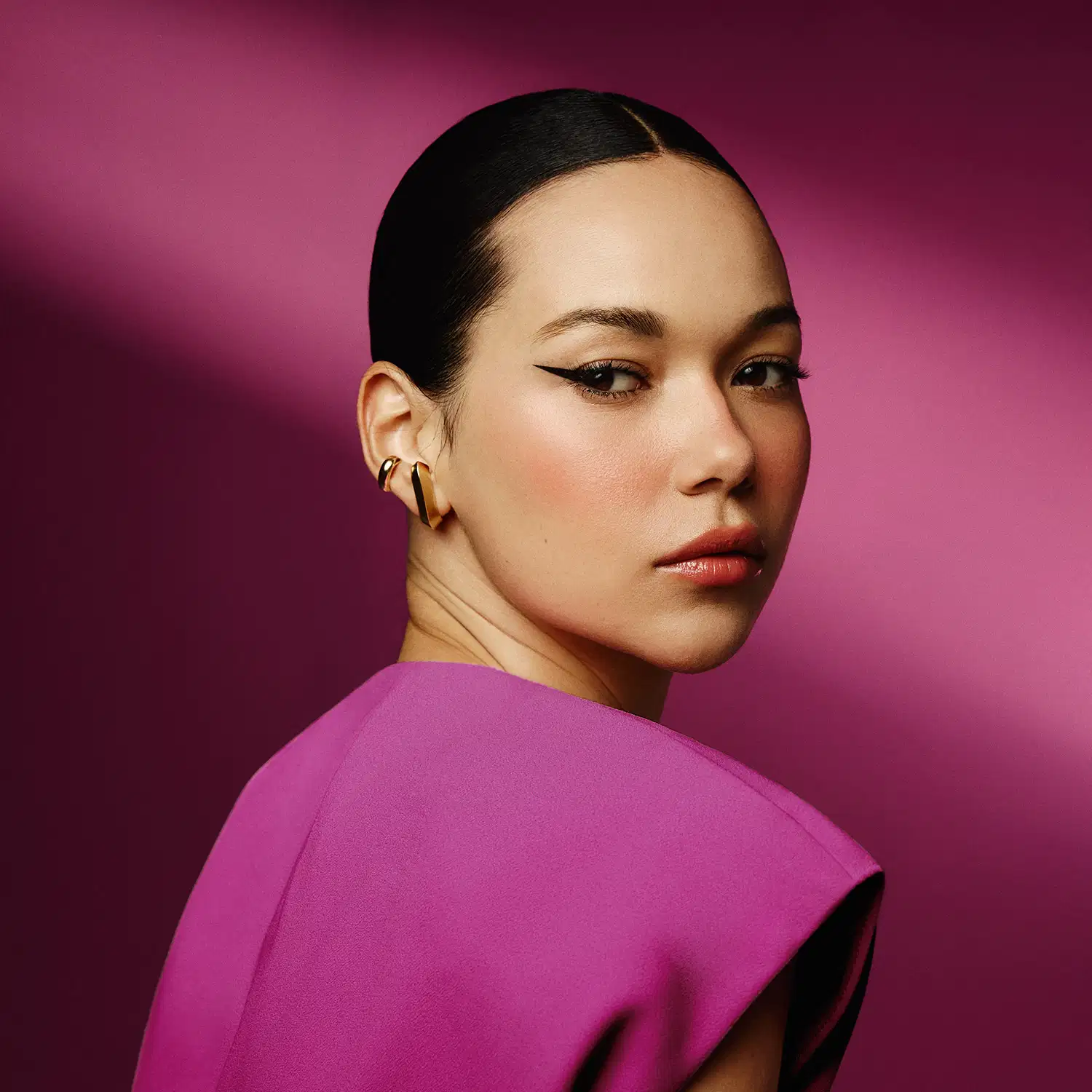 Ambiente de beleza elegante e sofisticado, com modelo feminino em destaque usando acessórios dourados e vestido rosa vibrante, em um cenário monocromático de fundo rosa intenso.