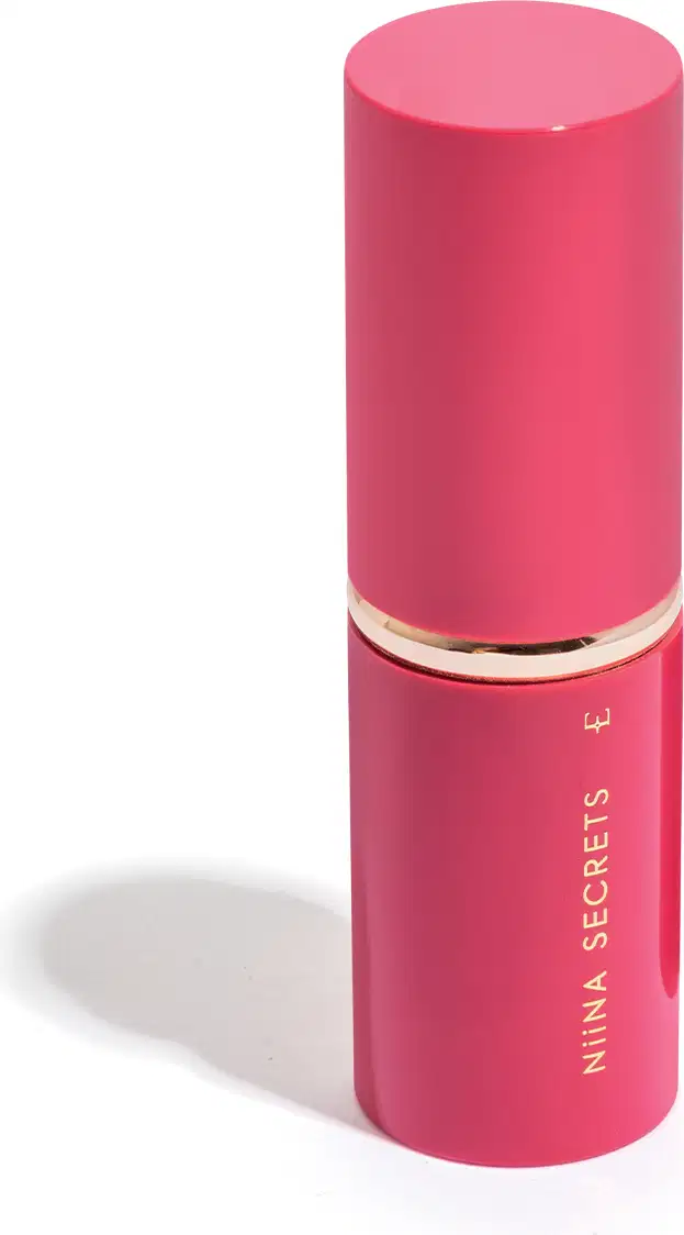 Recipiente cilíndrico rosa com tampa dourada. Rótulo com o texto "Nina Secrets" em letras douradas. Trata-se de um pincel retrátil com tampa.