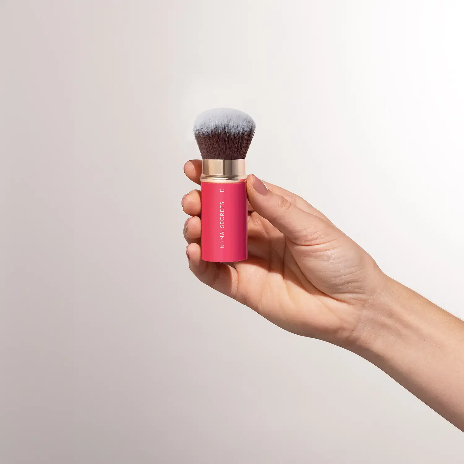 Uma mão segura o pincel de maquiagem retrátil com embalagem rosa. O item possui a marca "Nina Secrets" impressa em letras douradas.