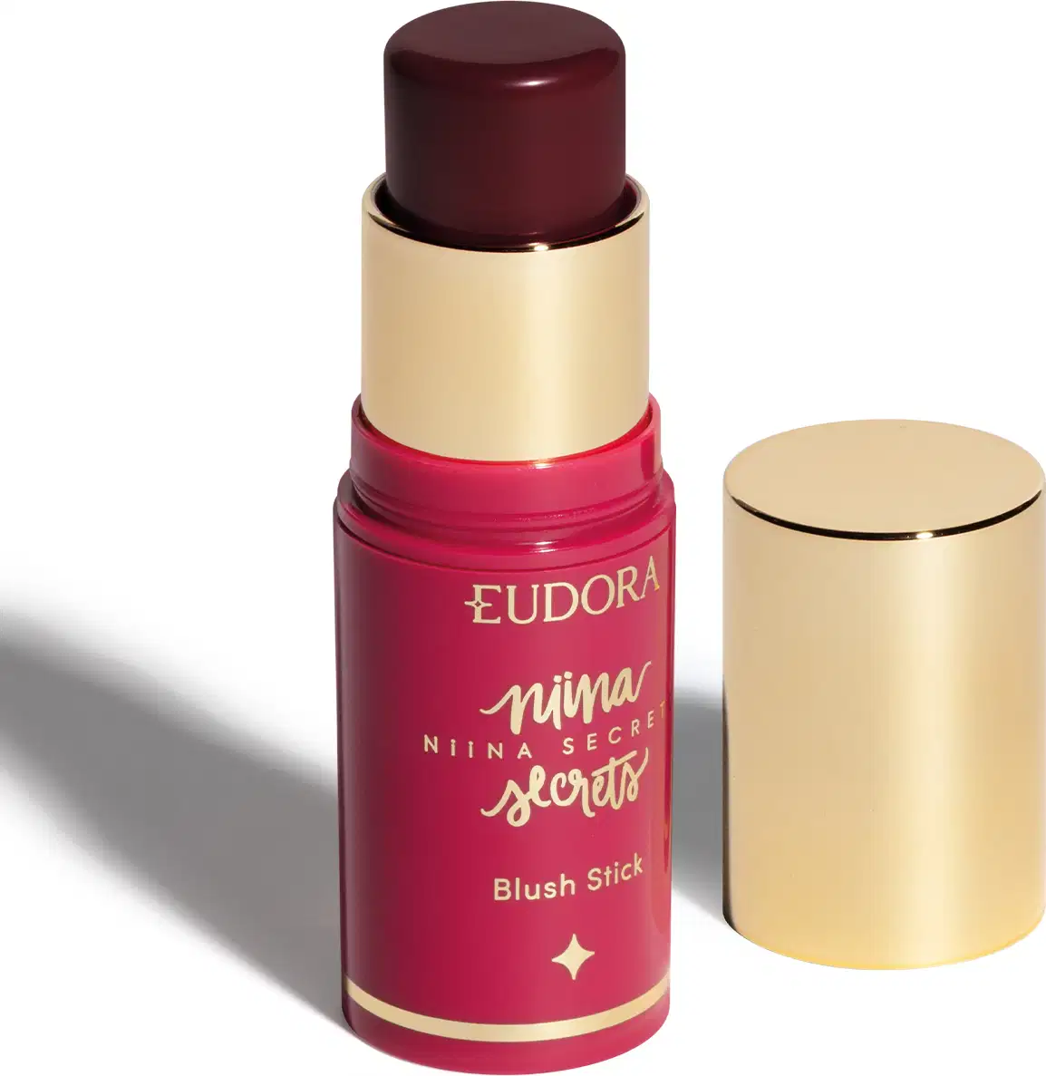 O produto apresentado é um blush em bastão da linha Niina Secrets da marca Eudora. A embalagem é composta por um tubo rosa com detalhes dourados. O produto está aberto, mostrando a cor Amora, com a tampa dourada ao lado.