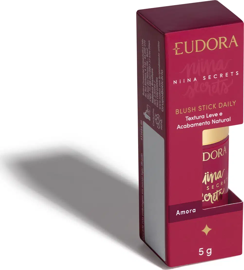 O produto apresentado é um blush stick da linha Niina Secrets da marca Eudora. A embalagem é uma caixa retangular de cor rosa com detalhes em dourado, contendo o nome da marca e da linha de produtos.