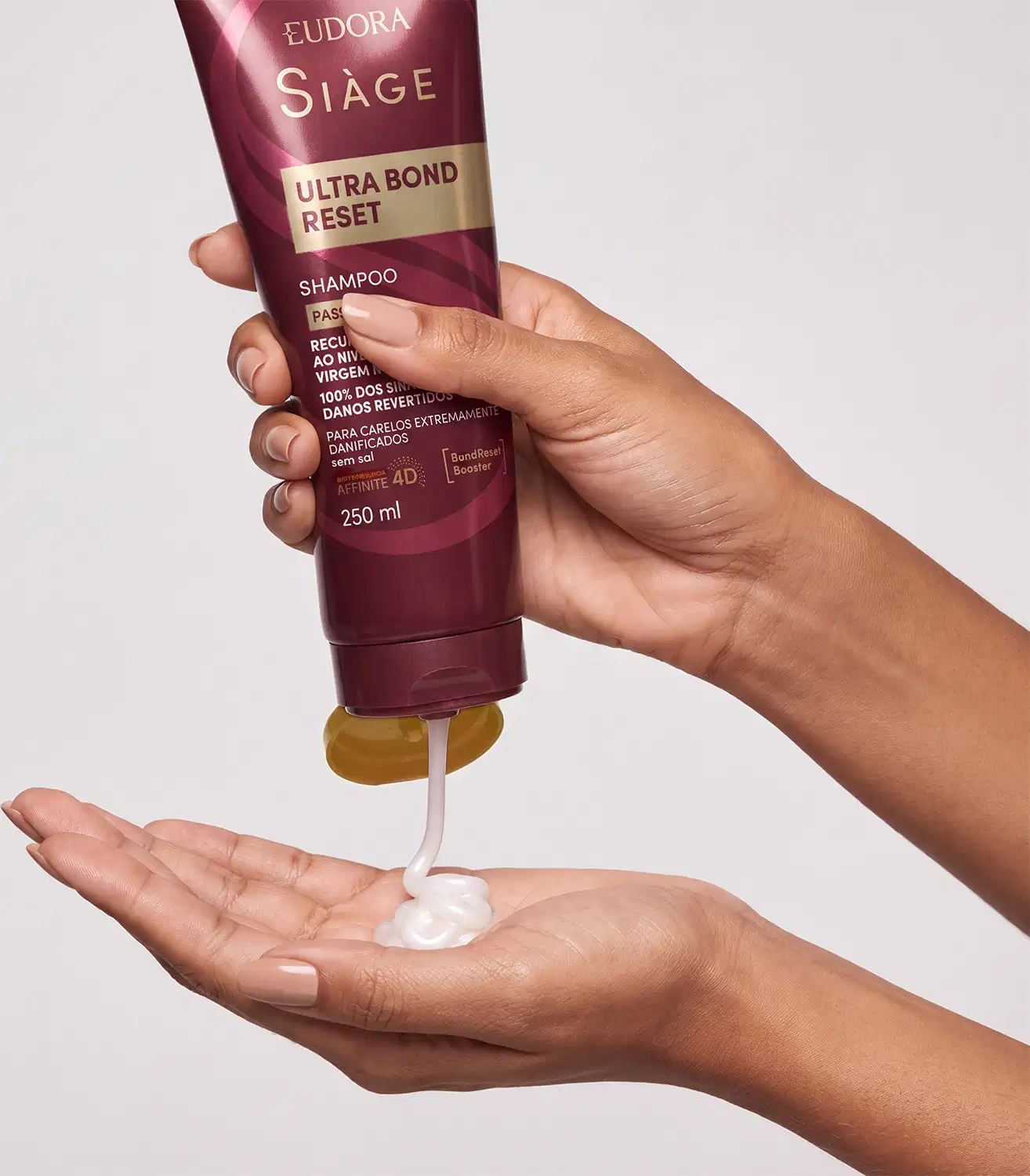 Tubo de shampoo para cabelos da marca Siàge, em tons de vinho e dourado. Uma mão está apertando a bisnaga e está saindo o produto, com textura cremosa e brilhante.