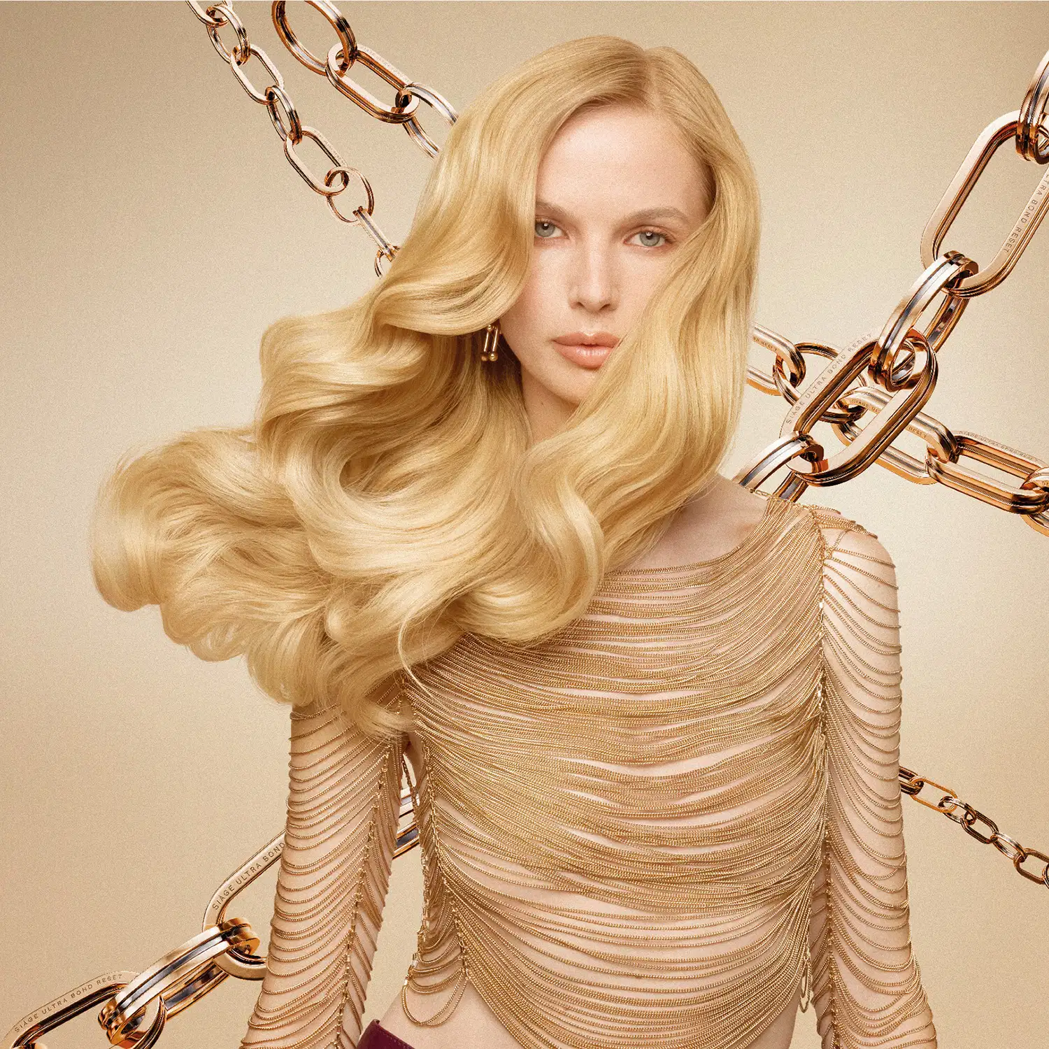 Uma modelo loira com cabelos longos e ondulados usa um vestido dourado com detalhes em correntes, em um cenário com fundo bege e elementos dourados, como correntes e anéis.