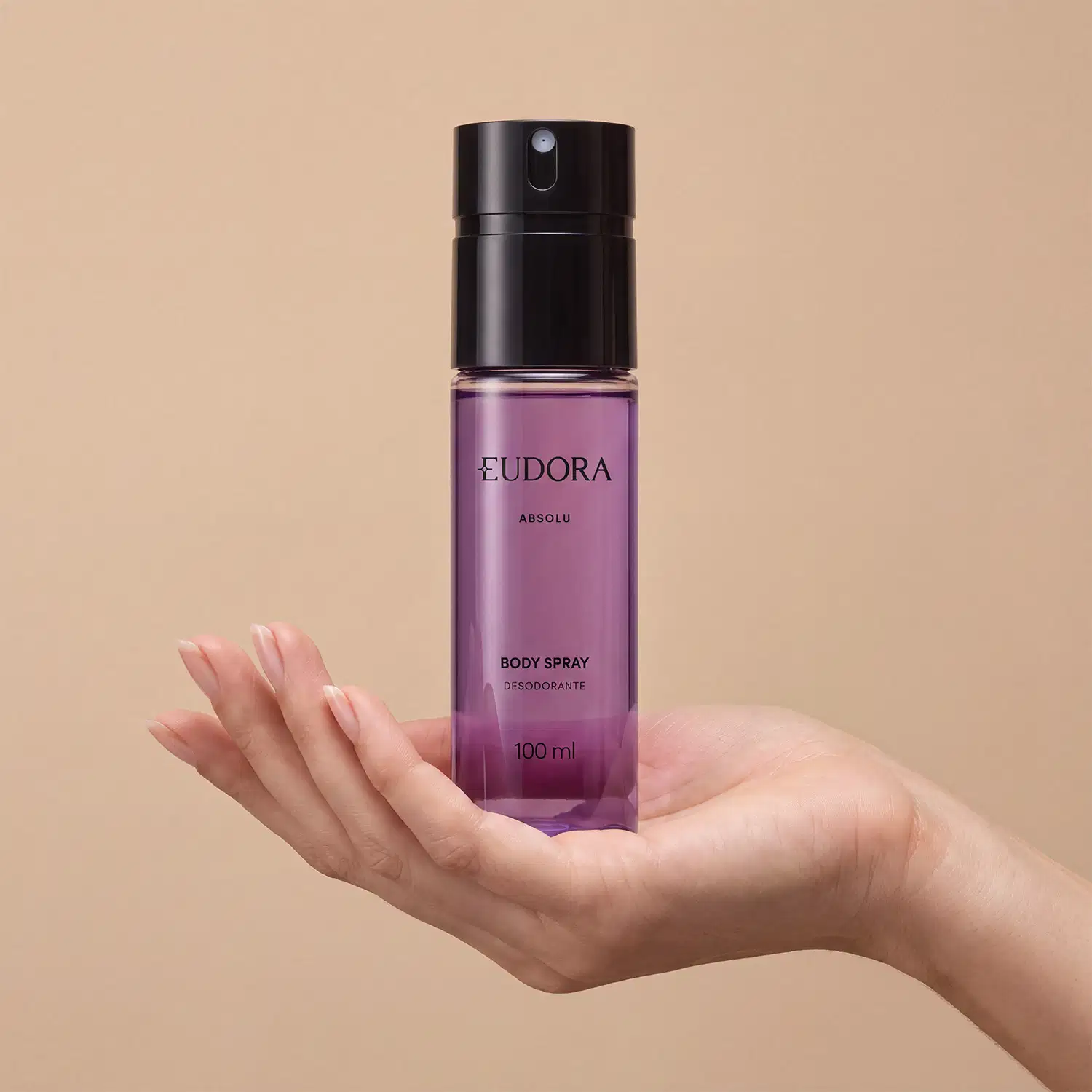 Frasco preto de item de perfumaria da marca Eudora, com líquido roxo claro, escrito: ABSOLU BODY SPRAY DESODORANTE em letras pretas. O produto está sendo segurado por mão feminina sobre fundo bege.
