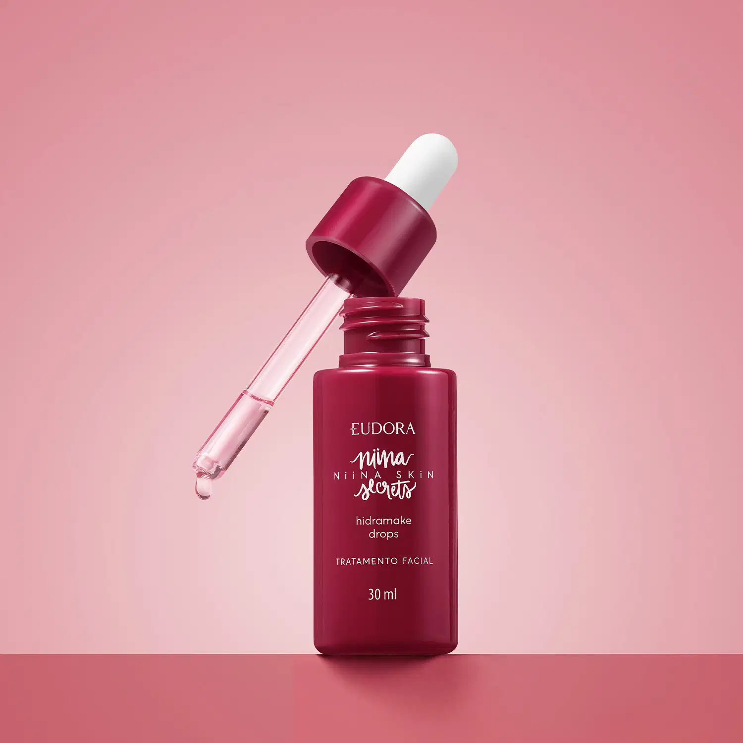 Frasco rosa com conta-gotas de sérum facial Eudora Niina Secrets Hidramake Drops 30ml, sobre fundo rosa com gotícula de produto visível no aplicador.