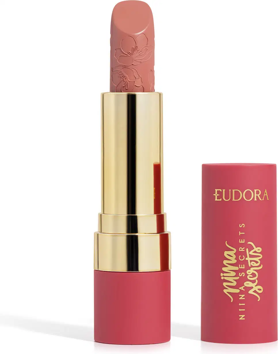 Embalagem de um batom aberto, com corpo dourado e tampa e base rosa, escrito "Eudora Niina Secrets". A cor do batom é rosé e tem um relevo exclusivo em padrão floral na bala. Produto de beleza com design elegante e sofisticado.

