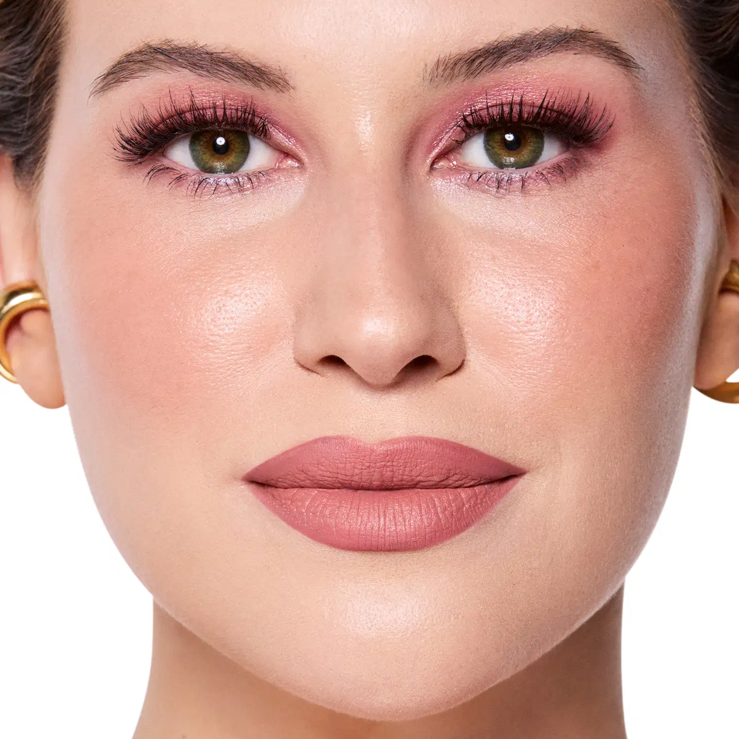 Rosto da Niina em foco, com maquiagem rosada e batom rosa claro semi-matte. Olhos verdes destacados por cílios longos e sobrancelhas bem definidas. Fundo claro e iluminado, sugerindo um ambiente de beleza ou cosmética.
