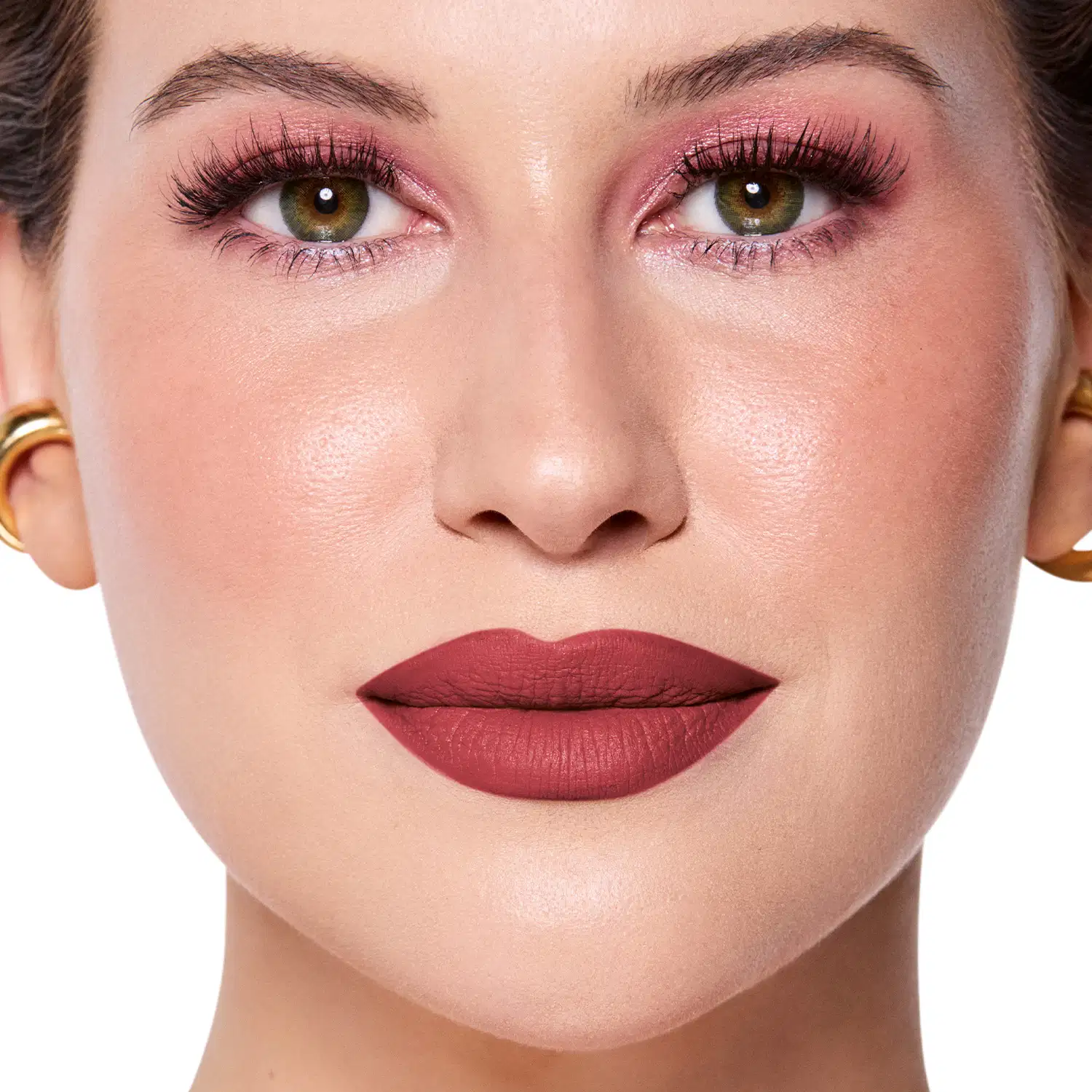Rosto da Niina em foco, com maquiagem rosada e batom vermelho semi-matte. Olhos verdes destacados por cílios longos e sobrancelhas bem definidas. Fundo claro e iluminado, sugerindo um ambiente de beleza ou cosmética.
