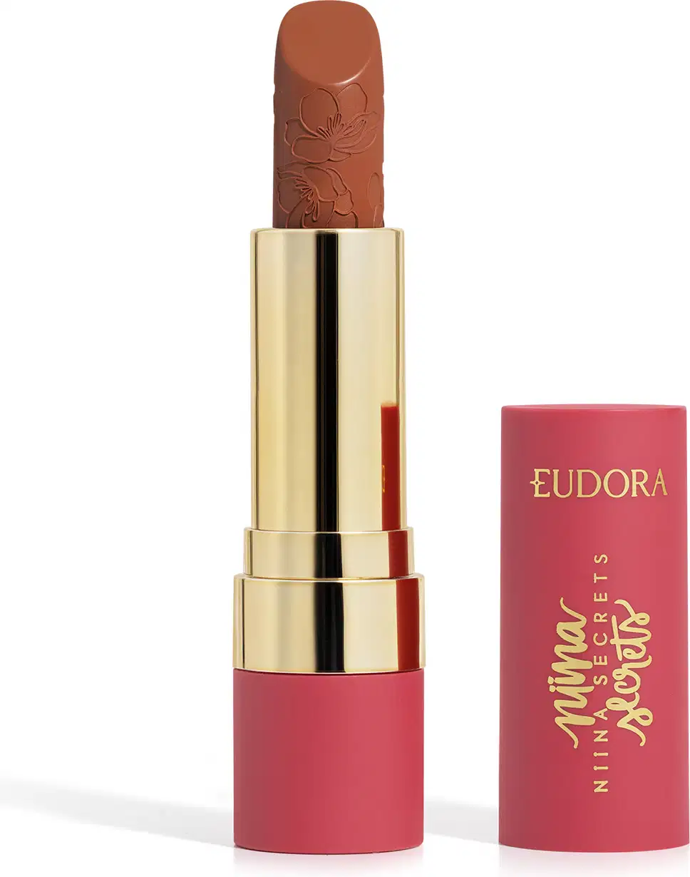Embalagem de um batom aberto, com corpo dourado e tampa e base rosa, escrito "Eudora Niina Secrets". A cor do batom é castanho e tem um relevo exclusivo em padrão floral na bala. Produto de beleza com design elegante e sofisticado.