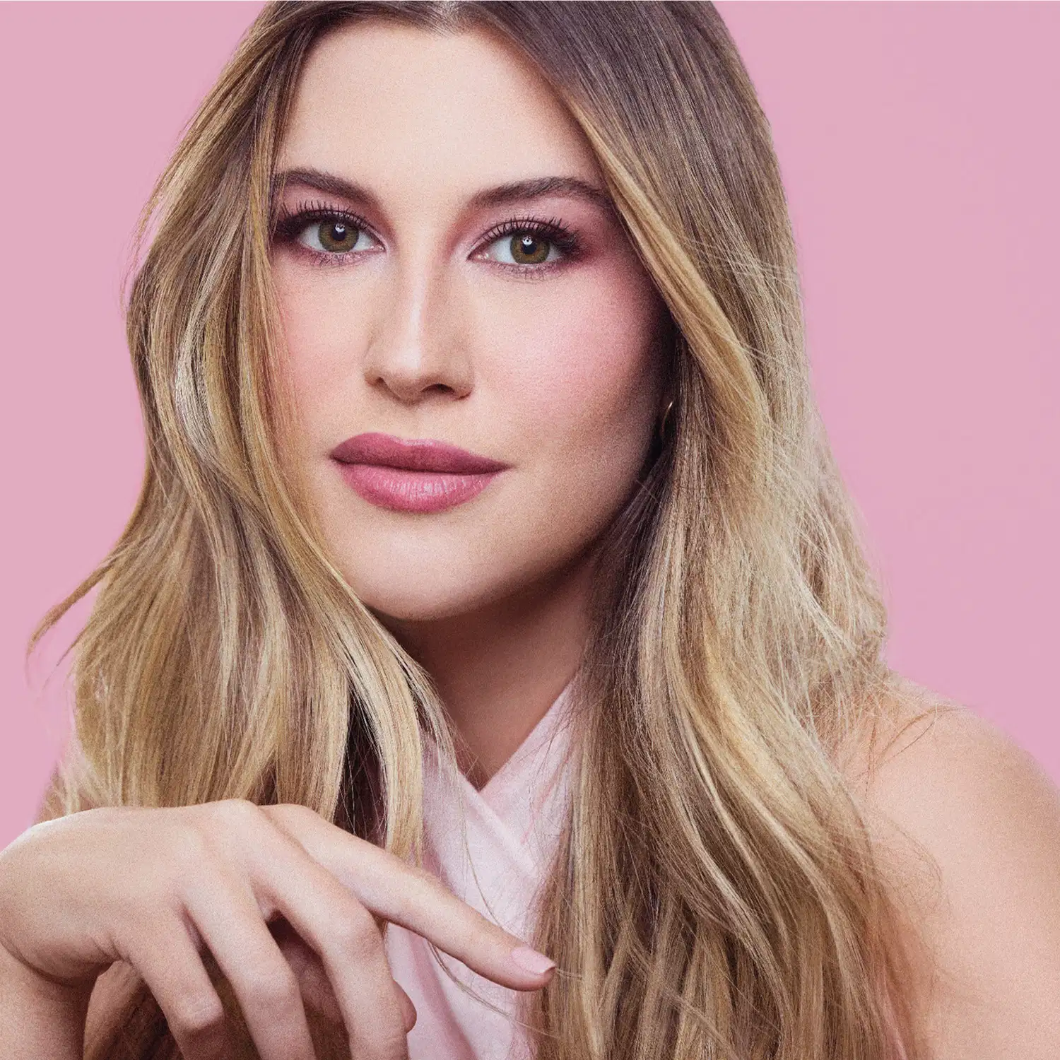 Mulher com cabelo louro comprido, maquiagem com sombra rosa e batom rosa, sobre fundo rosa uniforme, com ombro e braço visíveis.