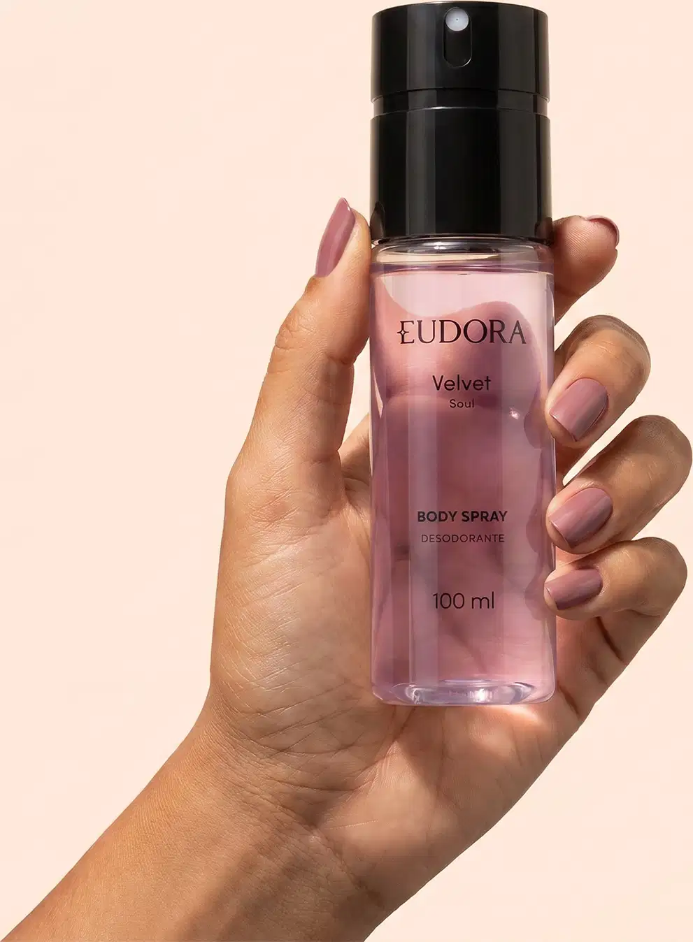 Mão segurando um frasco preto e roxo de item de perfumaria da marca Eudora, com rótulo "Velvet Soul" e informações sobre o produto. Fundo liso em tom de pêssego.