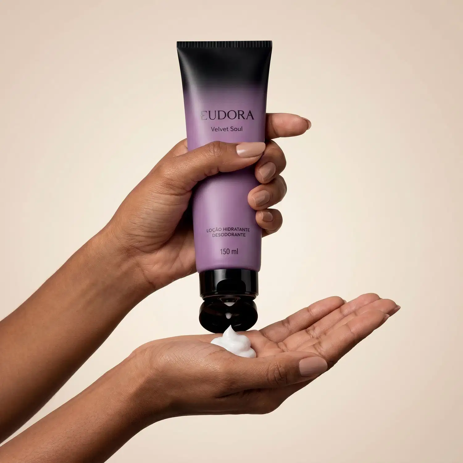 Tubo de loção hidratante desodorante da marca Eudora, com design em tons de roxo e preto. O tubo está sendo segurado por uma mão, e está saindo o produto com uma textura aveludada e aspecto cremoso, sugerindo uma consistência suave e agradável ao toque.