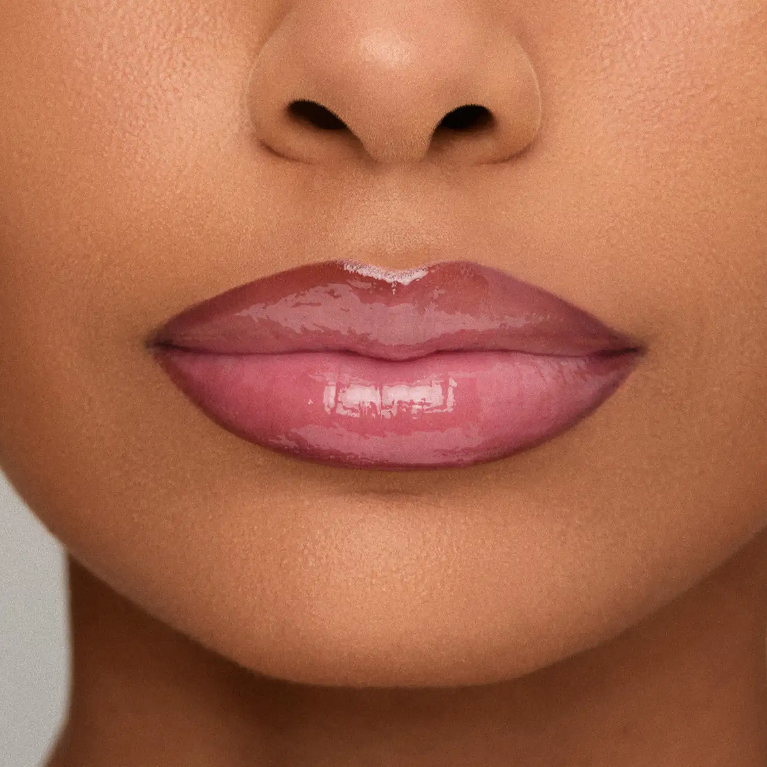 Imagem de close-up de lábios femininos com lapiseira labial e gloss amarronzado brilhante, em fundo claro e suave. 
