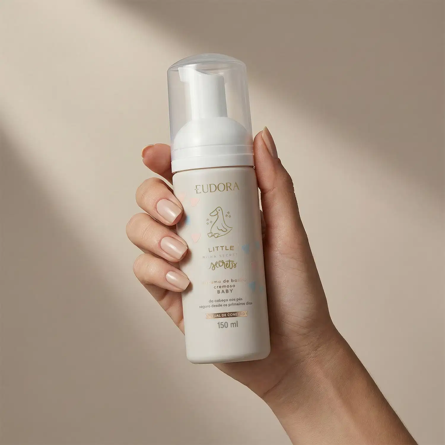 Imagem de um produto de cuidados para bebê da marca Eudora, com embalagem branca e detalhes em tons claro. Segurado por uma mão feminina, em um ambiente neutro e iluminado.