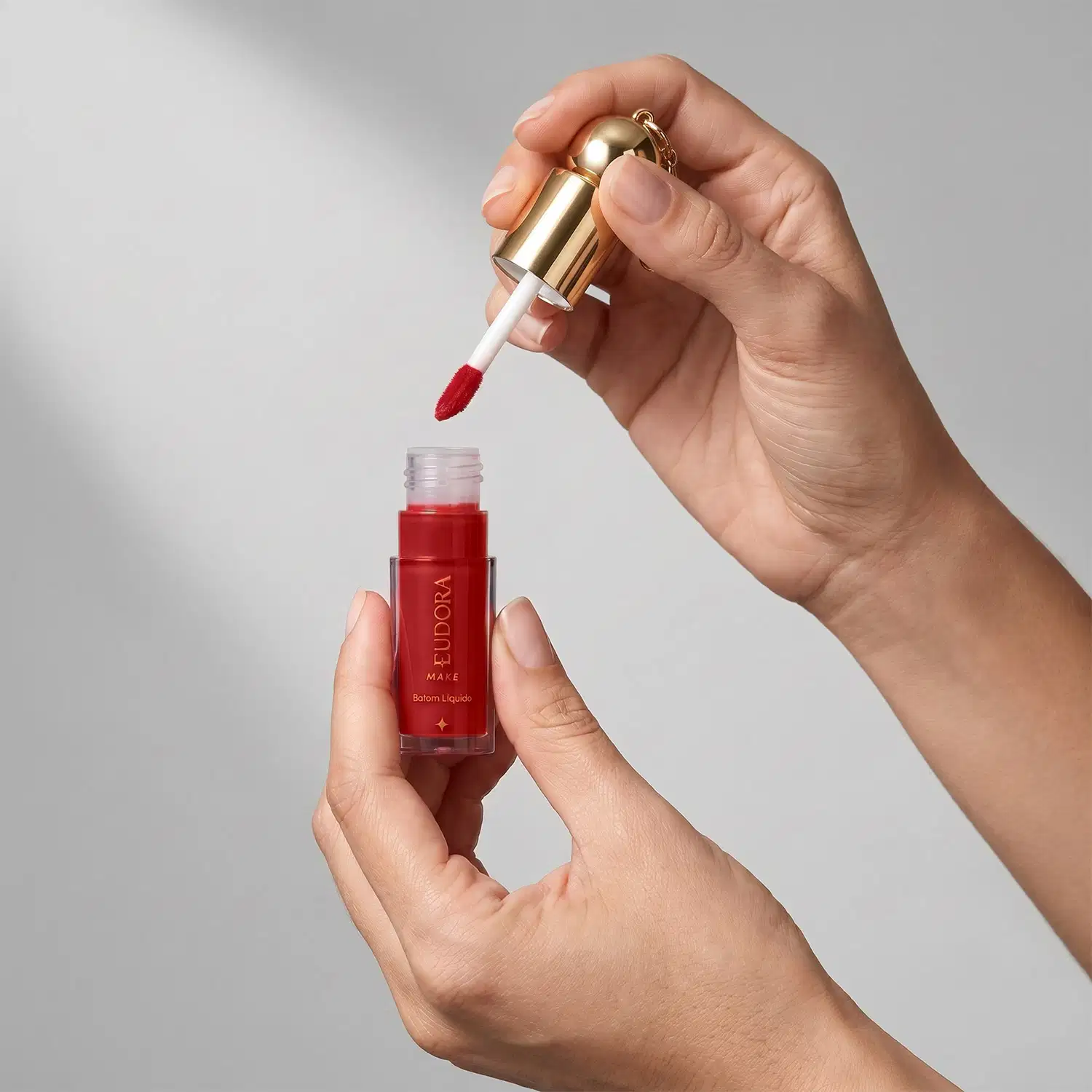 Mãos segurando o batom líquido aberto, com aplicador. O item está sobre um fundo cinza claro. A cor do produto é em tom de vermelho.
