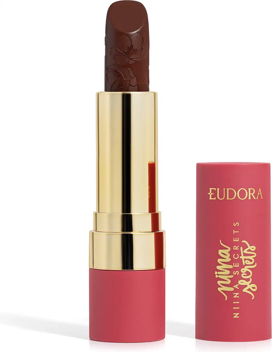 Embalagem de um batom aberto, com corpo dourado e tampa e base rosa, escrito "Eudora Niina Secrets". A cor do batom é marrom escuro e tem um relevo exclusivo em padrão floral na bala. Produto de beleza com design elegante e sofisticado.