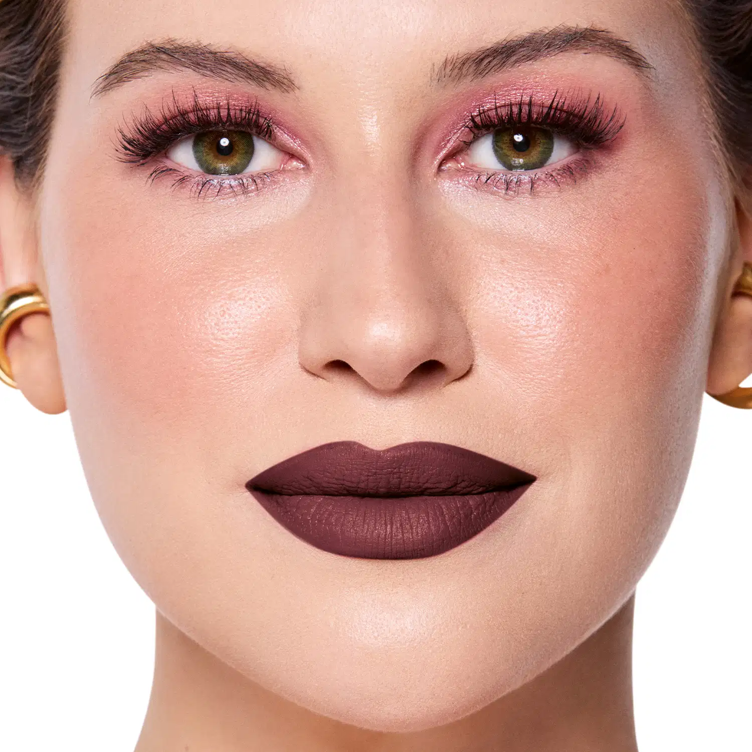 Rosto da Niina em foco, com maquiagem rosada e batom marrom escuro semi-matte. Olhos verdes destacados por cílios longos e sobrancelhas bem definidas. Fundo claro e iluminado, sugerindo um ambiente de beleza ou cosmética.
