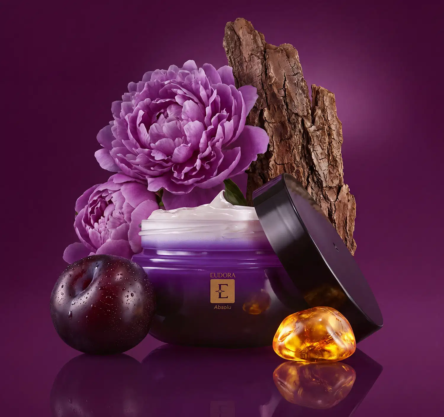 Recipiente de creme corporal roxo da marca Eudora, com flores de peônia, uma ameixa preta, pedaços de sândalo, e pedra de âmbar dourado, indicando os ingredientes que fazem parte da família olfativa. Sobre fundo roxo escuro.