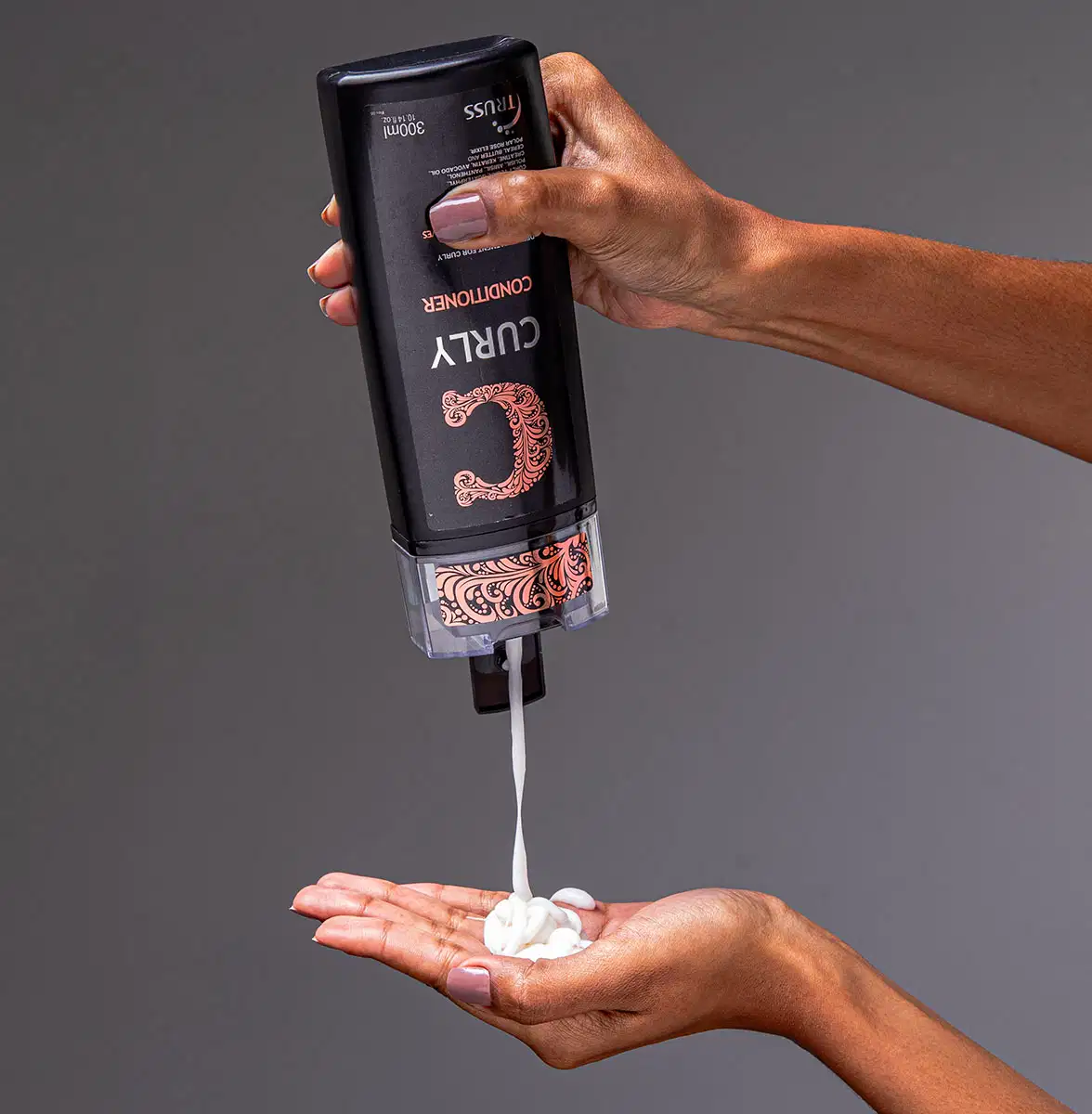 A imagem mostra uma mão segurando um frasco de condicionador da marca Truss, linha Curly. Também é possível ver o seu conteúdo, que é cremoso e branco, com 300ml.