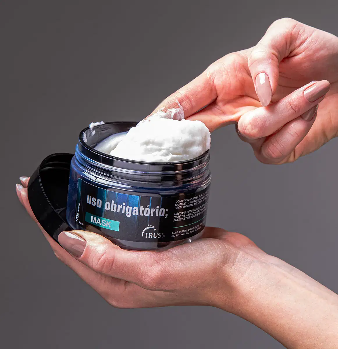 A imagem mostra uma mão está segurando o pote da máscara capilar e aplicando o produto branco cremoso com os dedos.