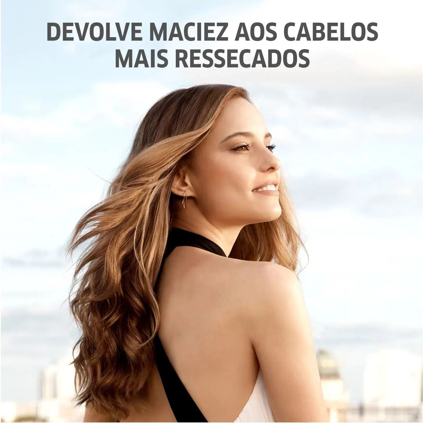 A imagem apresenta uma mulher jovem com longos cabelos ondulados em tons de loiro e castanho. Ela está usando um top preto e tem um sorriso suave no rosto. O texto diz "Devolve maciez aos cabelos mais ressecados".