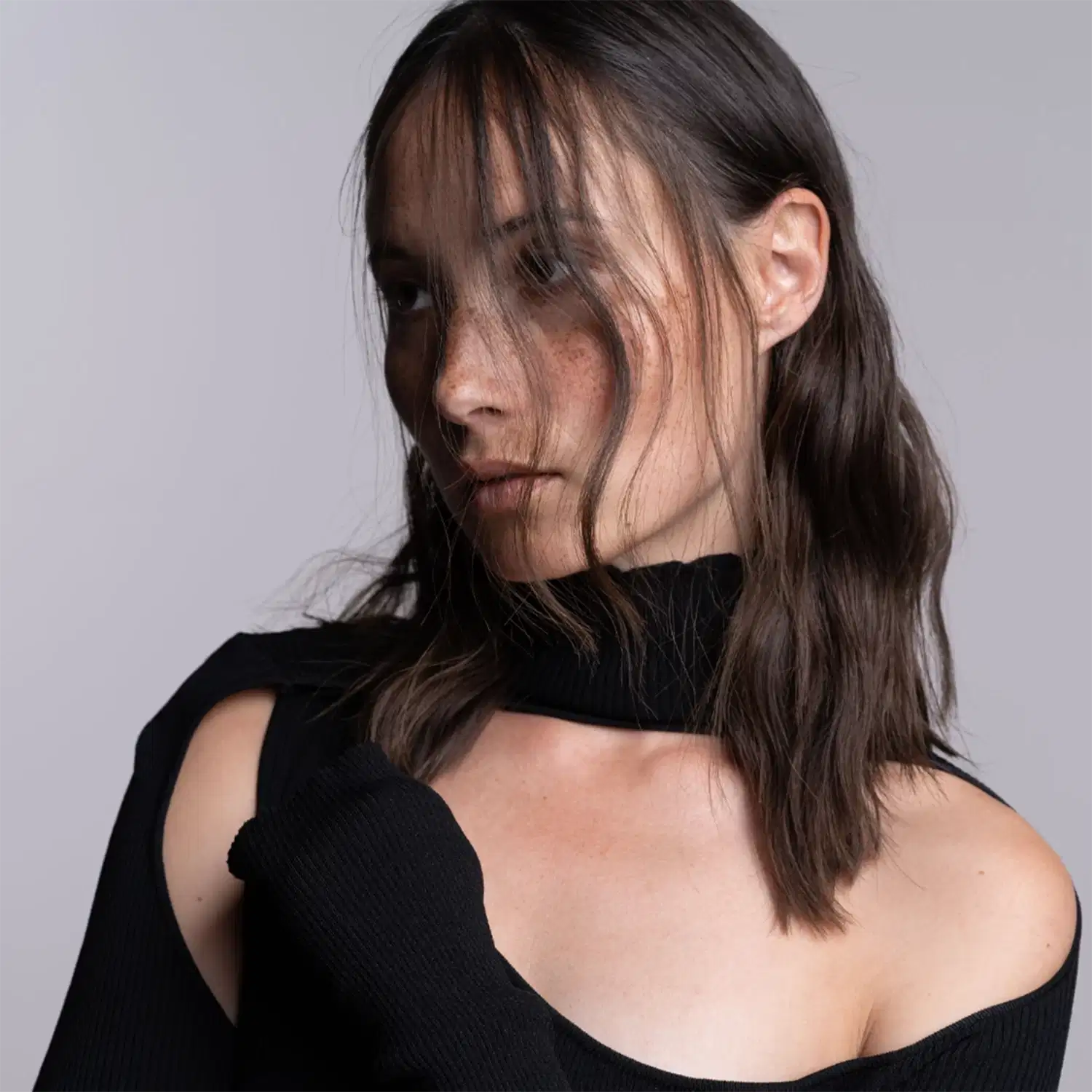 Esta imagem mostra uma mulher com longos cabelos castanhos caindo sobre seu rosto. Ela está usando uma blusa preta sem mangas que deixa seus ombros à mostra. A imagem é um close-up que foca no rosto e ombros da mulher contra um fundo neutro e desfocado.