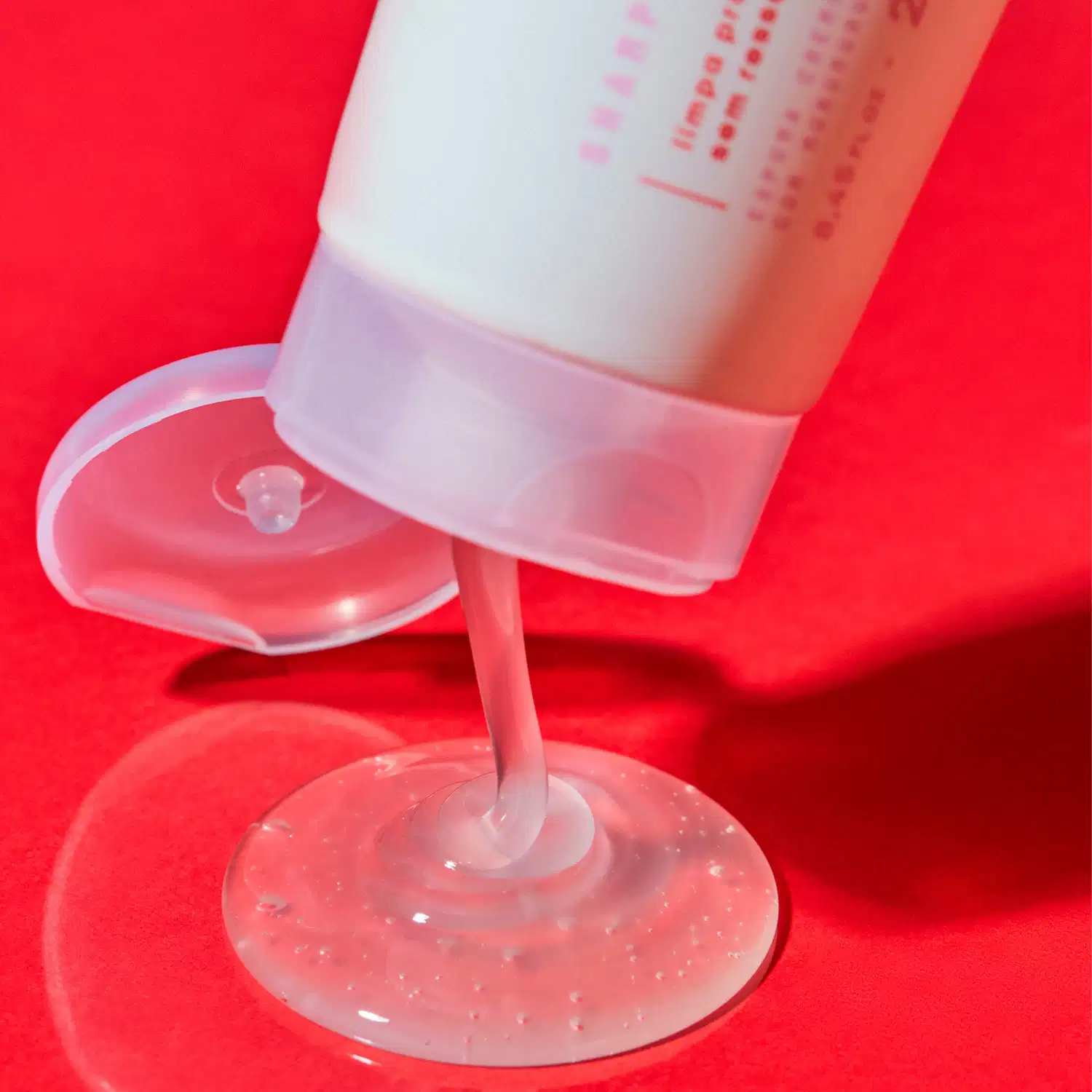 Gel ou sérum translúcido em frasco com aplicador de disco de silicone, sendo despejado sobre superfície vermelha. Embalagem branca com rótulo rosa e texto em português.