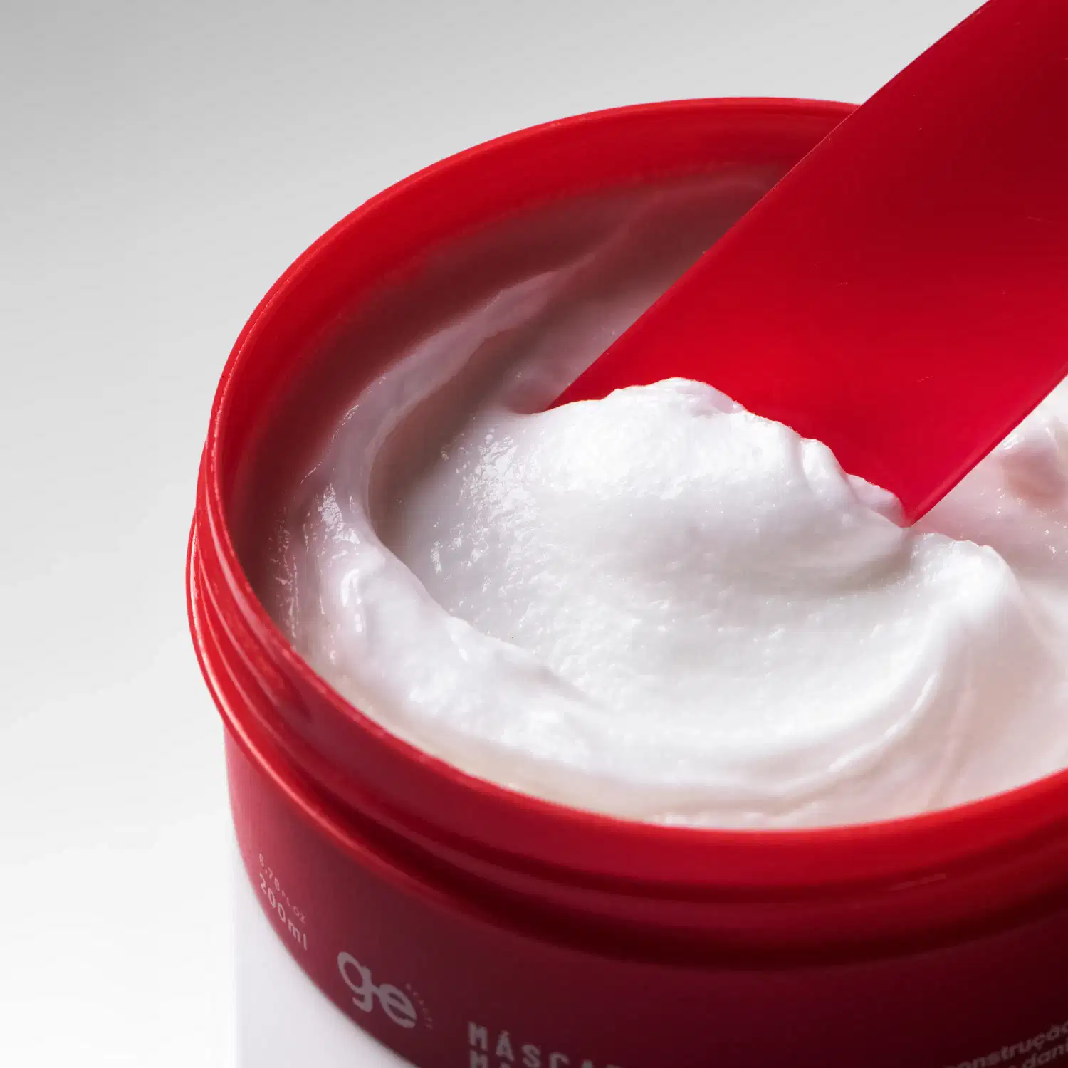 Creme branco, denso e levemente brilhante, de aparência cremosa e macia, com consistência espessa que forma picos ao ser espalhado com a espátula vermelha.
