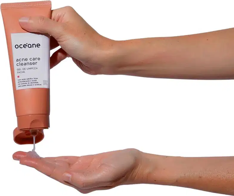 A imagem mostra um par de mãos segurando a embalagem do Océane Acne Care Cleanser. Uma das mãos está apertando o tubo para dispensar o produto líquido na outra mão aberta.
