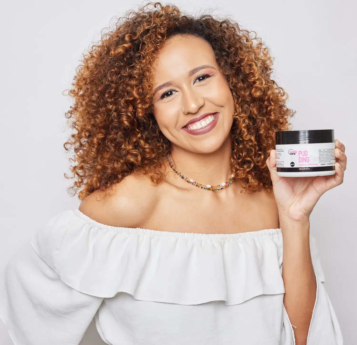 Pote plástico branco com tampa preta contendo um produto de definição de cachos da marca Curly Care. O rótulo indica que é um "Pudding Gelatina Definidora" vegano, com pH 4,2.