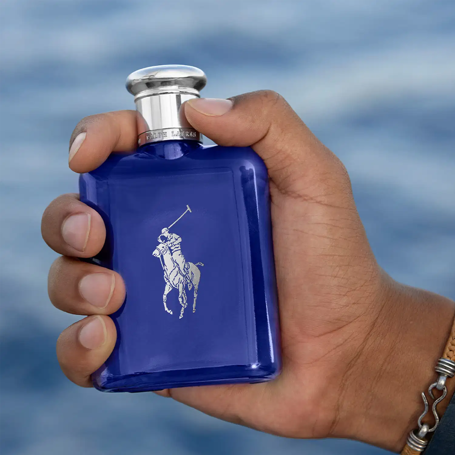 Perfume em frasco retangular de vidro azul cobalto com o logotipo do cavalo empinado branco da Ralph Lauren. O frasco tem uma tampa cromada arredondada. O produto está sendo segurado por uma mão masculina com pulseira de metal prateada, contra um céu.