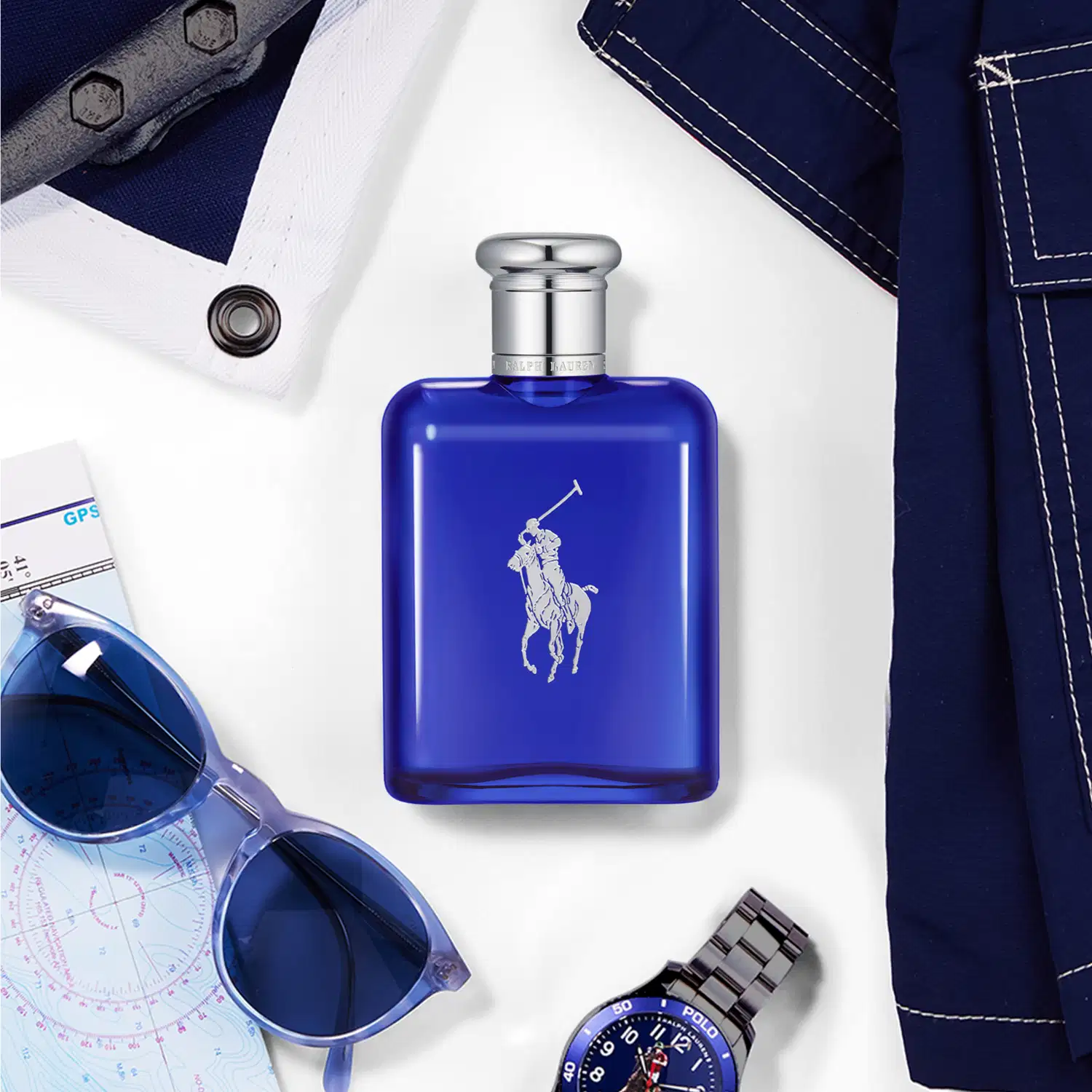 Perfume em frasco retangular de vidro azul escuro com logotipo do cavalo e cavaleiro e tampa prateada. O frasco está acompanhado por óculos de sol azuis, relógio esportivo azul marinho, mapa náutico e peças de vestuário jeans.